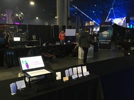 A photo of a laptop and some booklets set up at a convention, with lights from a stage and people visible in the background.