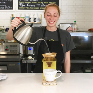 A photo of a smiling person pouring water into a pour-over coffee set-up.