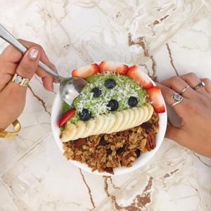 A photo of somebody's hands holding and digging a spoon into a green smoothie bowl decorated with fruits and granola.